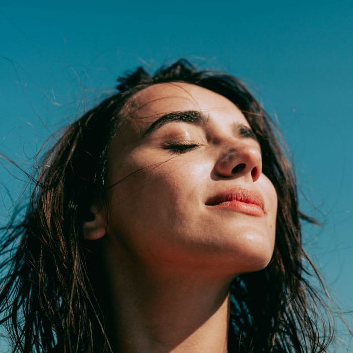 Femme fermant les yeux en souriant. Grand ciel bleu en fond. La femme est face au soleil comme pour se ressourcer en vitamines, les cheveux au vent. Cette image fait référence au patch énergie